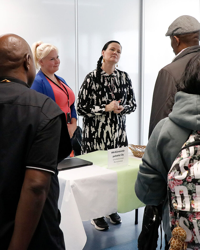 Kirjavamekkoinen henkilö keskustelee hattupäisen henkilön kanssa.