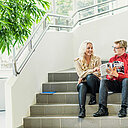 Kaksi ihmistä istuu TAKKin Tampereen valtatien kampuksen aulan rappusilla ja selaa koulutusopasta.