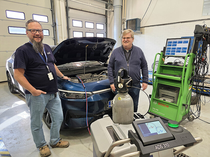 Kouluttajat Mika Sivenius ja Juha Vainio seisovat Tampereen Aikuiskoulutuskeskuksen autohallissa, taustalla auto konepelti auki. Autoon on kytketty hiilidioksidi-ilmastoinninhuoltolaite.