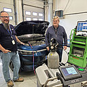 Kouluttajat Mika Sivenius ja Juha Vainio seisovat Tampereen Aikuiskoulutuskeskuksen autohallissa, taustalla auto konepelti auki. Autoon on kytketty hiilidioksidi-ilmastoinninhuoltolaite.