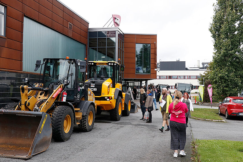 Kiinteistönvälitysalan työkoneet jonoon ajettuna Nirvan kampuksen edustalla, vieressä kävelijöitä. 