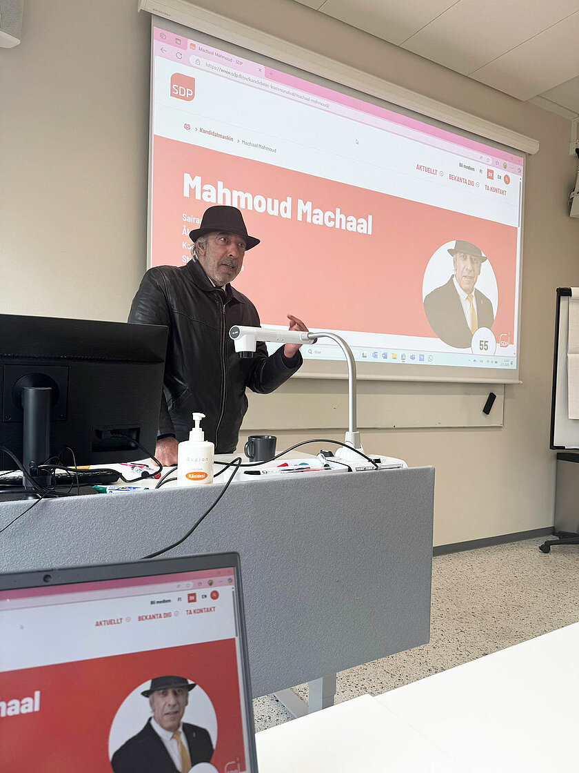 Hattupäinen mies puhumassa pöydän takana. 