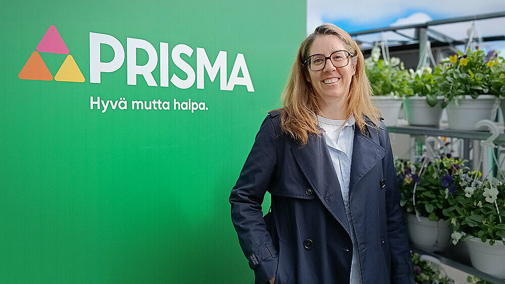 Nainen hymyilee iloisesti Prisma-logon edessä kukkia taustalla.
