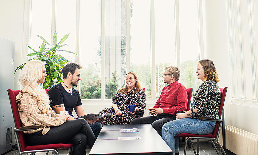 Ihmisiä kahvilla matalan pöydän ääressä, taustalla korkea ikkuna, josta tulvii valoa.