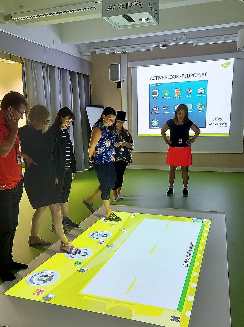 Neljä pelaajaa pelaamassa tietovisaa. Kaksi henkilöä seuraa tilannetta taustalla.