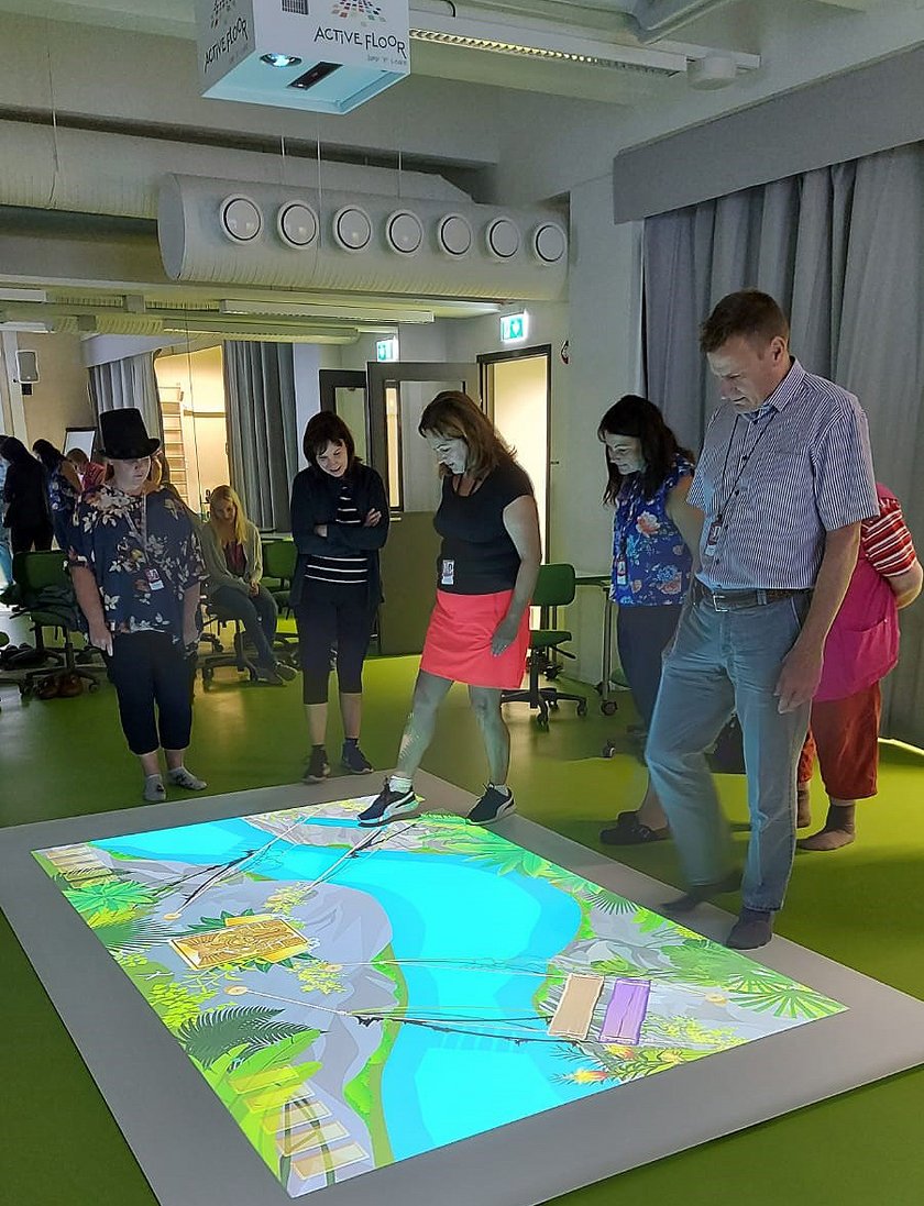 Kaksi henkilöä testaa Aktiivilattian viidakkoseikkailu-tietopeliä. Viisi henkilöä seuraa tilannetta.