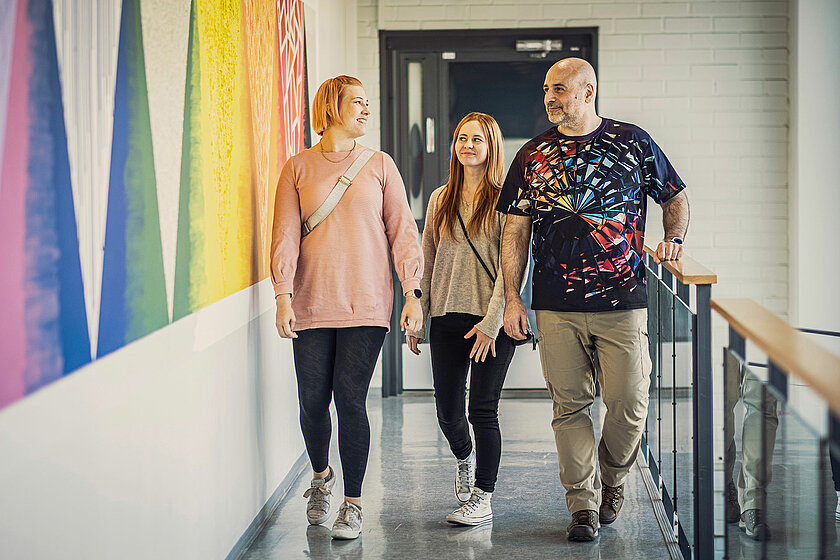 Kolme henkilöä kävelee rinnakkain valoisalla käytävällä. 