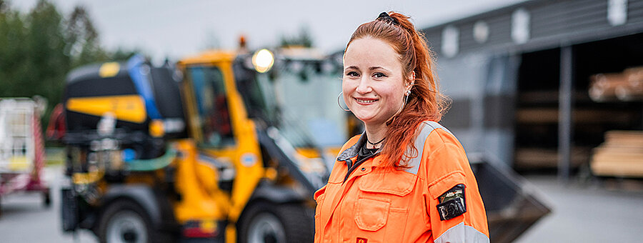Nainen oransseissa työvaatteissa keltaisen kiinteistöpalvelutyökoneen edessä.