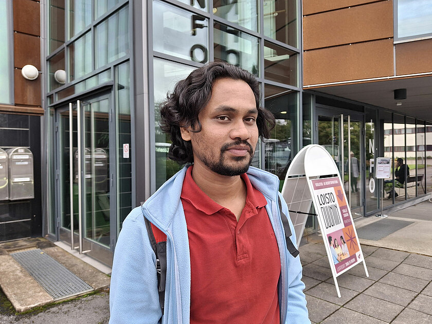 Punapaitainen Erfan seisoo TAKKin Nirvan kampuksen infonedustalla, taustalla näkyvät infon lasiseinät. 