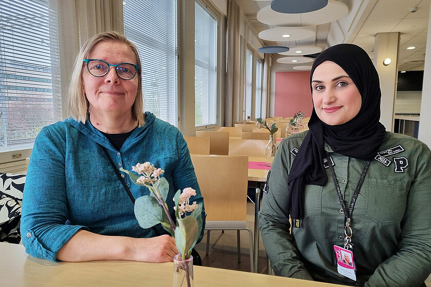 Anukaisa ja Samah istuvat pöydän ääressä, taustalla ikkunarivistö, edessä kukka. 