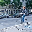 Mies ajaa kadulla pyörällä, jossa on valtava etupyörä ja pieni takapyörä.
