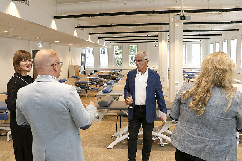 Taustalla näkyy urheiluhieronnan luokka. Etualalla ihmiset keskustelevat juhlavaatteet ylllään.