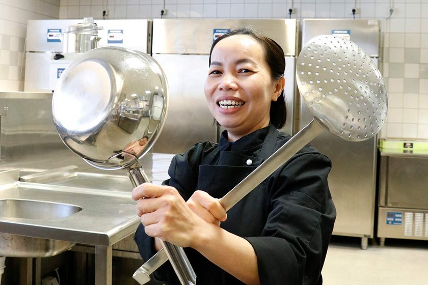 A woman is holding two ketteles and smiling in cook's black outfit..