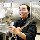 A woman is holding two ketteles and smiling in cook's black outfit..