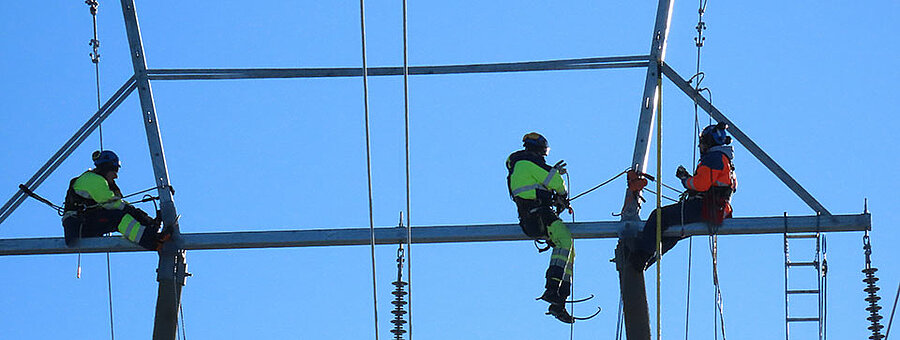 Kolme henkilöä istuu korkealla voimajohdon päällä vaihtamassa eristintä.