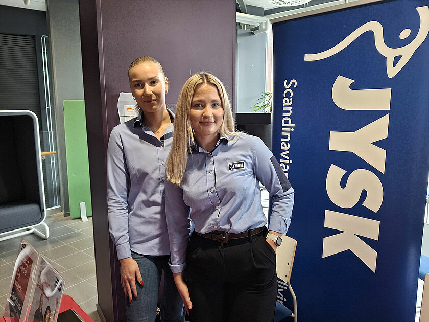 Kaksi vaaleansiniseen kauluspaitaan pukeutunutta henkilöä seisoo JYSK-kyltin edessä. 