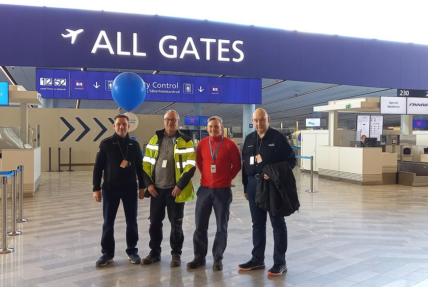 Mikko Mänty, Timo Ahonen, Keijo Halonen ja Heikki Mahlamäki Helsinki-Vantaan lentoasemalla. Katon rajassa suuri sininen kyltti, joka johdattaa lähtöporteille. 