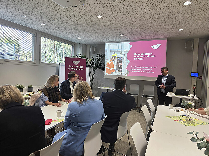 Rehtori Jarno Tuimala pitämässä PowerPoint-esitystä, pöydässä ihmiset istuvat ja kuuntelevat.