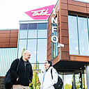 Henkilöt seisovat oppilaitoksen rakennuksen edustalla, takana näkyvät rakennuksen suuret lasi-ikkunat ja TAKKin logo.