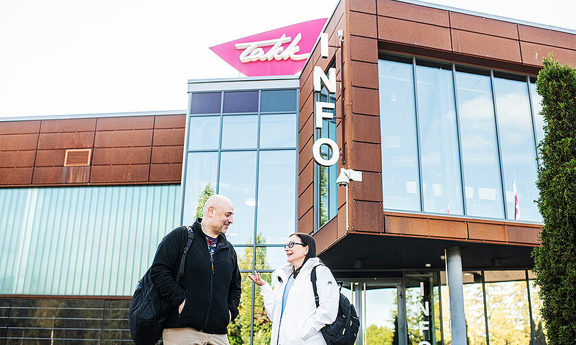 Henkilöt seisovat oppilaitoksen rakennuksen edustalla, takana näkyvät rakennuksen suuret lasi-ikkunat ja TAKKin logo.