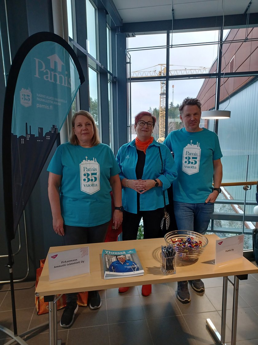 Pamislaiset seisovat kapean ja puisen esittelypöydän takana, ikkunoiden edessä. Pöydällä lehtiä, kyniä ja karkkia.