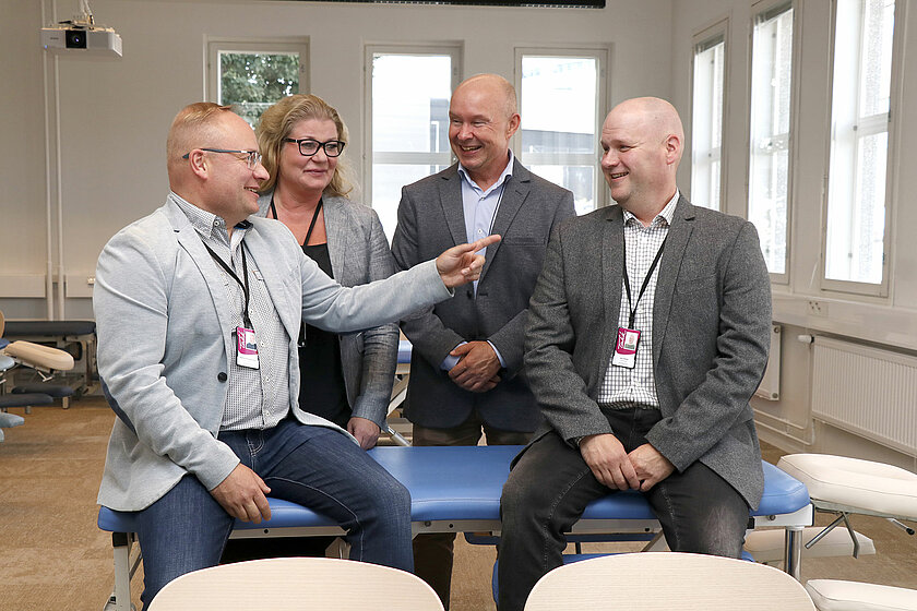 Urheiluhieronnan kouluttajat iloitsemassa avajaisissa, kaksi istuu hierontapöydällä ja kaksi seisoo heidän takanaan.