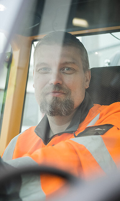 Henkilö työkoneen ratissa oranssissa työvaatteessa.