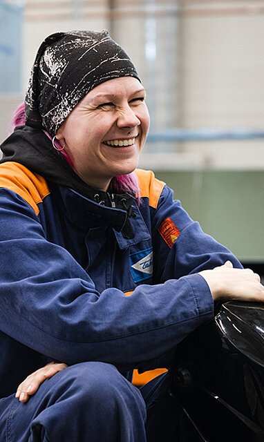 Hitsaajanainen istuu työhaalarit päällä hitsauskypärä kädessä TAKKin hitsaushallissa.