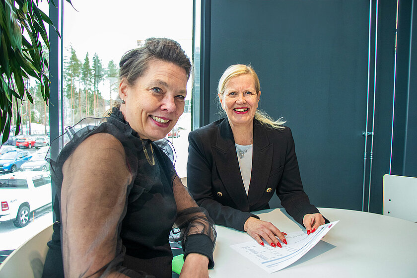 Kaksi naista istuu pöydän ääressä keskustelemassa. Taustalla suuri ikkuna, josta näkyy parkkipaikka.