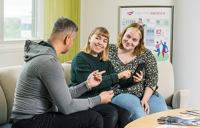 Kolme opiskelijaa istuu sohvalla, yhdellä puhelin kädessä ja näyttää sitä muille. 