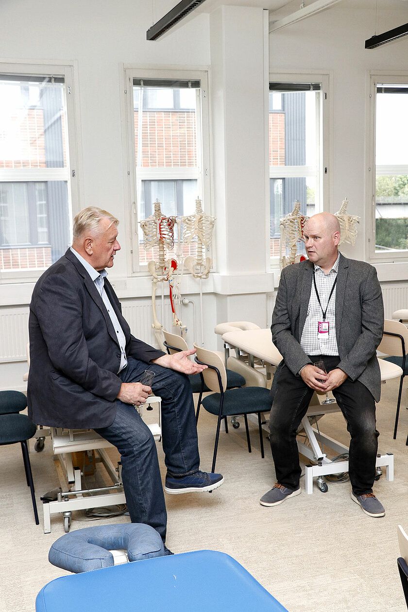 Kaksi henkilöä istuu hierontapöydillä ja keskustelee. 