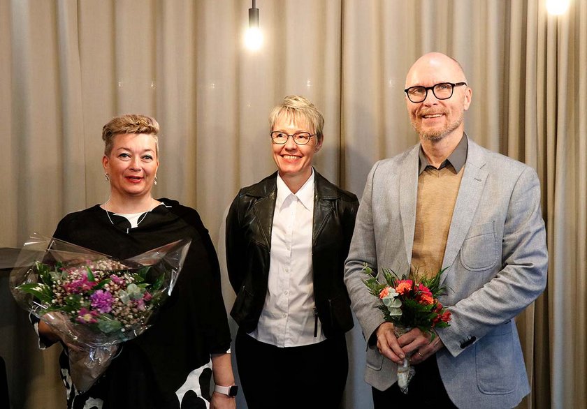 Johanna Toivanen, Kirsi Vuori ja Arto Kauppinen hymyilevät harmaan verhon edessä, käsissä kukkia.