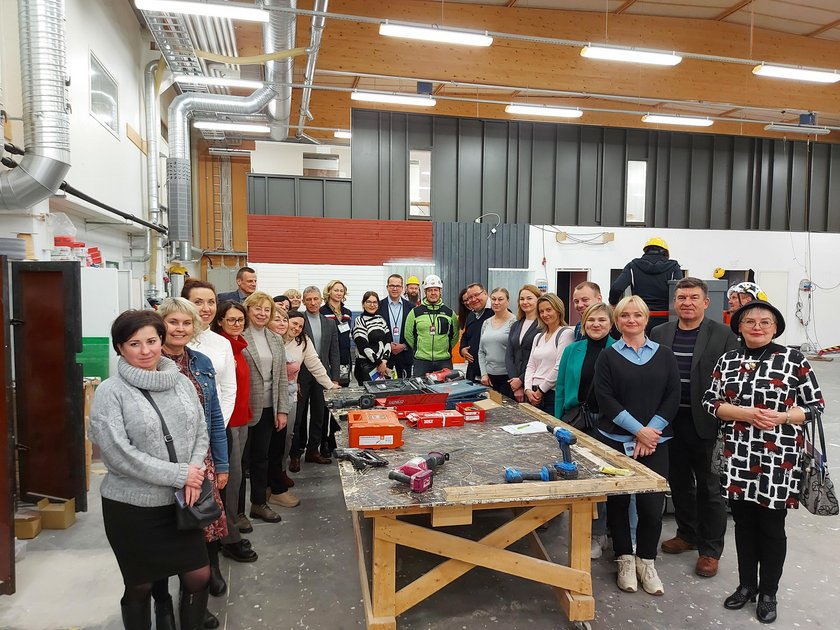 20 ihmistä seisomassa ison pöydän ympärillä rakennusalan harjoitteluhallissa.
