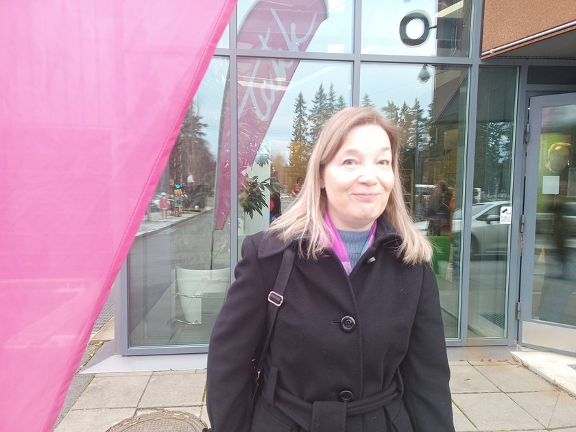 Tuija seisoo ulkona TAKKin infon ikkunoiden edessä laukku olallaan ja hymyilee.