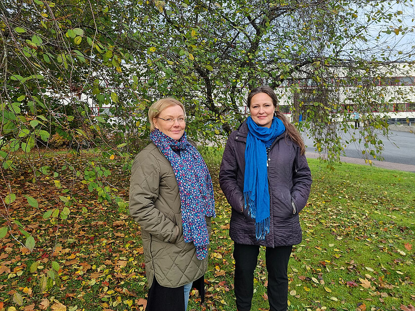 Hanna ja Ilse seisovat kädet taskuissa syksyisen puun alla ja hymyilevät. 
