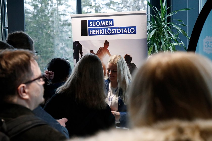 Vaaleahiuksinen henkilö seisoo Suomen Henkilöstötalo -mainosseinän edessä, pisteen edessä paljon työnhakijoita.