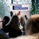 Vaaleahiuksinen henkilö seisoo Suomen Henkilöstötalo -mainosseinän edessä, pisteen edessä paljon työnhakijoita.