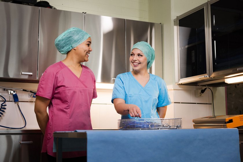 Kaksi työasuun pukeutunutta välinehuoltajaa sinisellä liinalla peitetyn pöydän ääressä. Pöydällä instrumentteja.