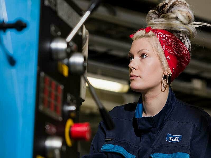 Elina Jaakola sorvin ääressä Nainen sinisissä työhaalareissa koneistuskoneen ääressä.