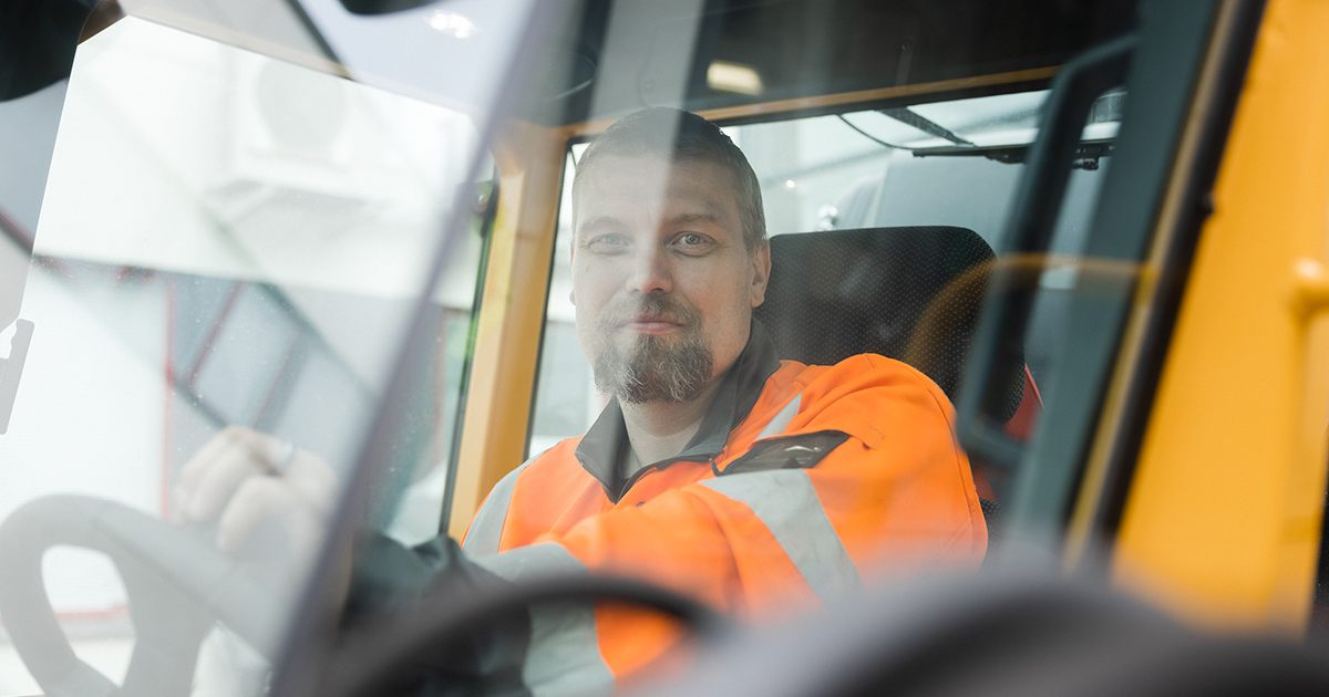 Henkilö työkoneen ratissa oranssissa työvaatteessa.