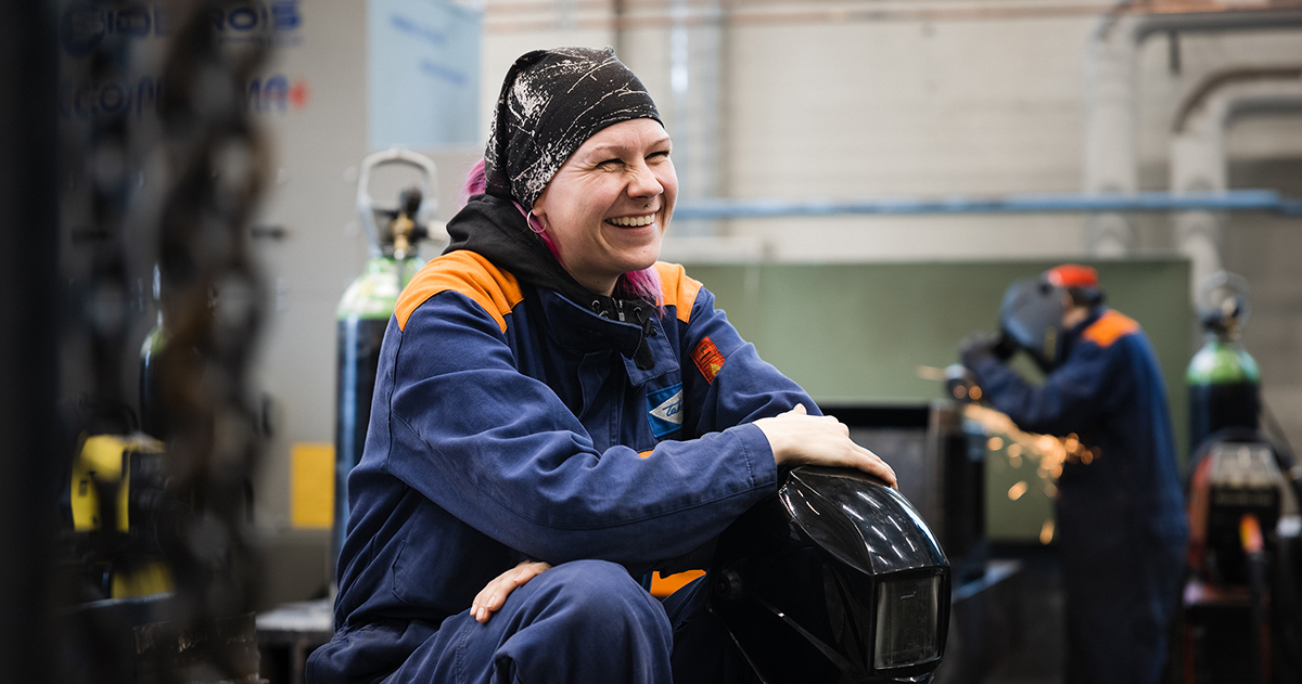 Hitsaajanainen istuu työhaalarit päällä hitsauskypärä kädessä TAKKin hitsaushallissa.