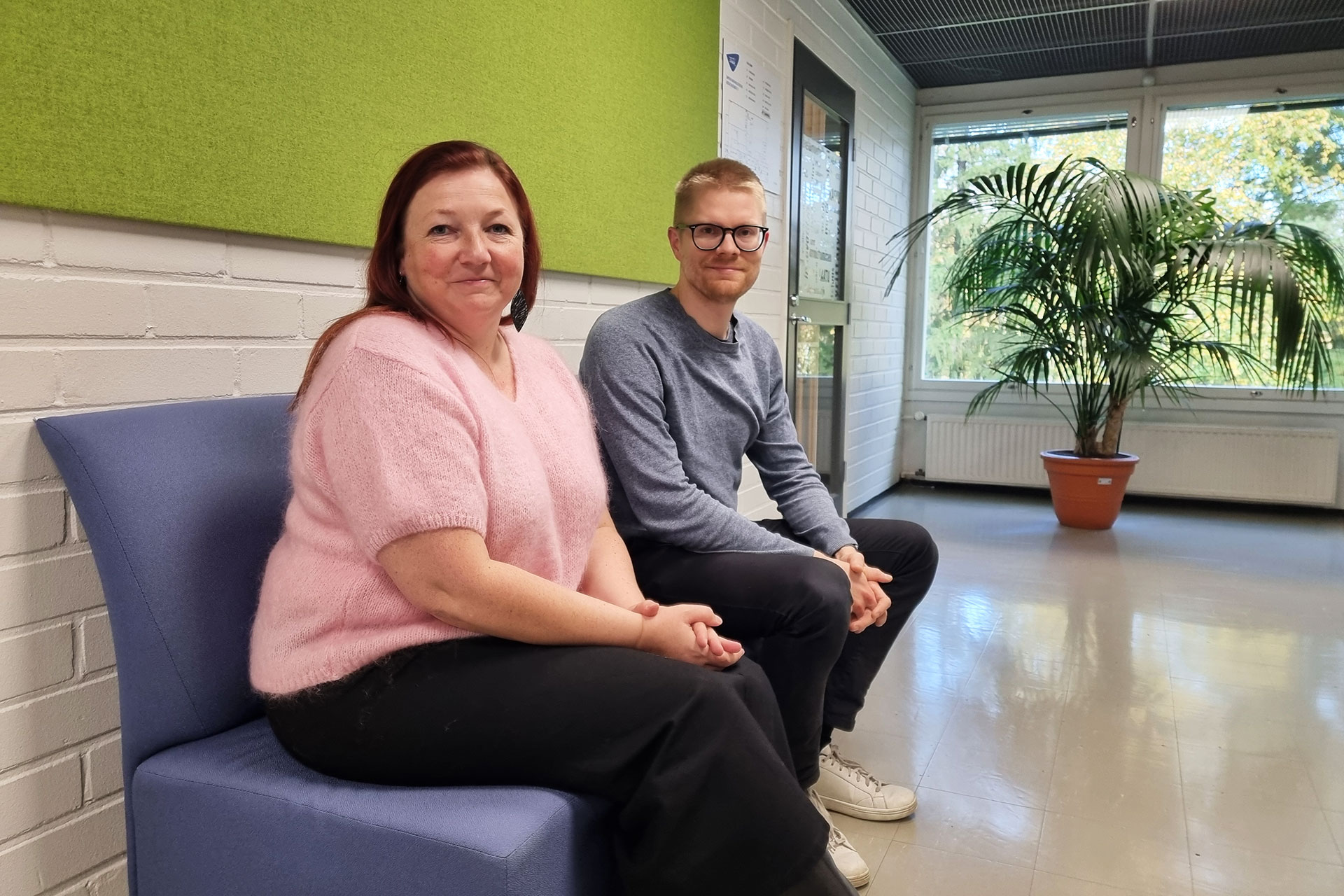 Kaksi henkilöä istuu käytävällä sinisellä sohvalla, takana vihreä seinä. Käytävän päässä ikkuna ja viherkasvi.