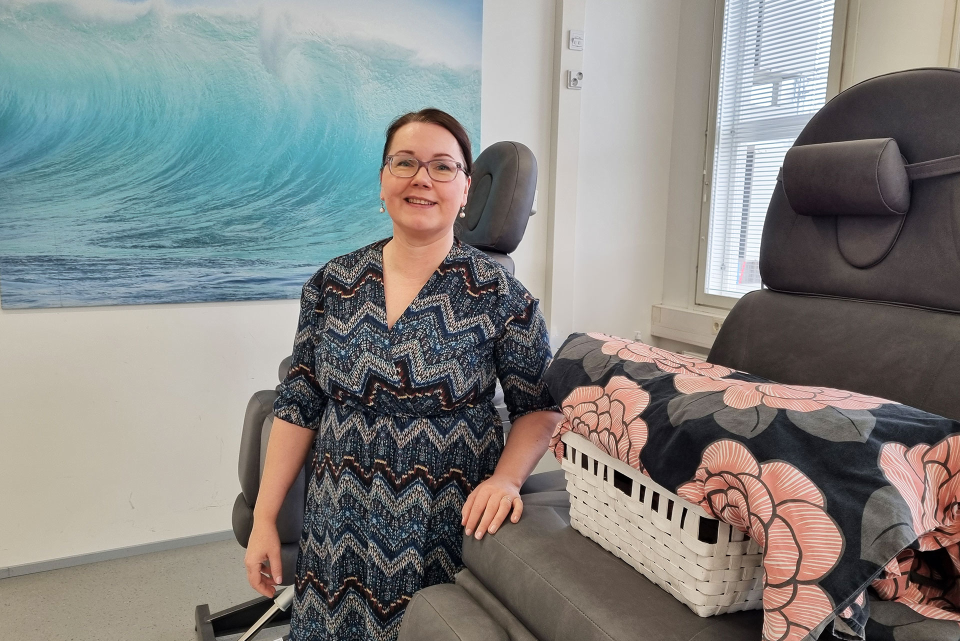 Katri seisoo jalkojenhoitotuolin vieressä, taustalla turkoosi taulu. 