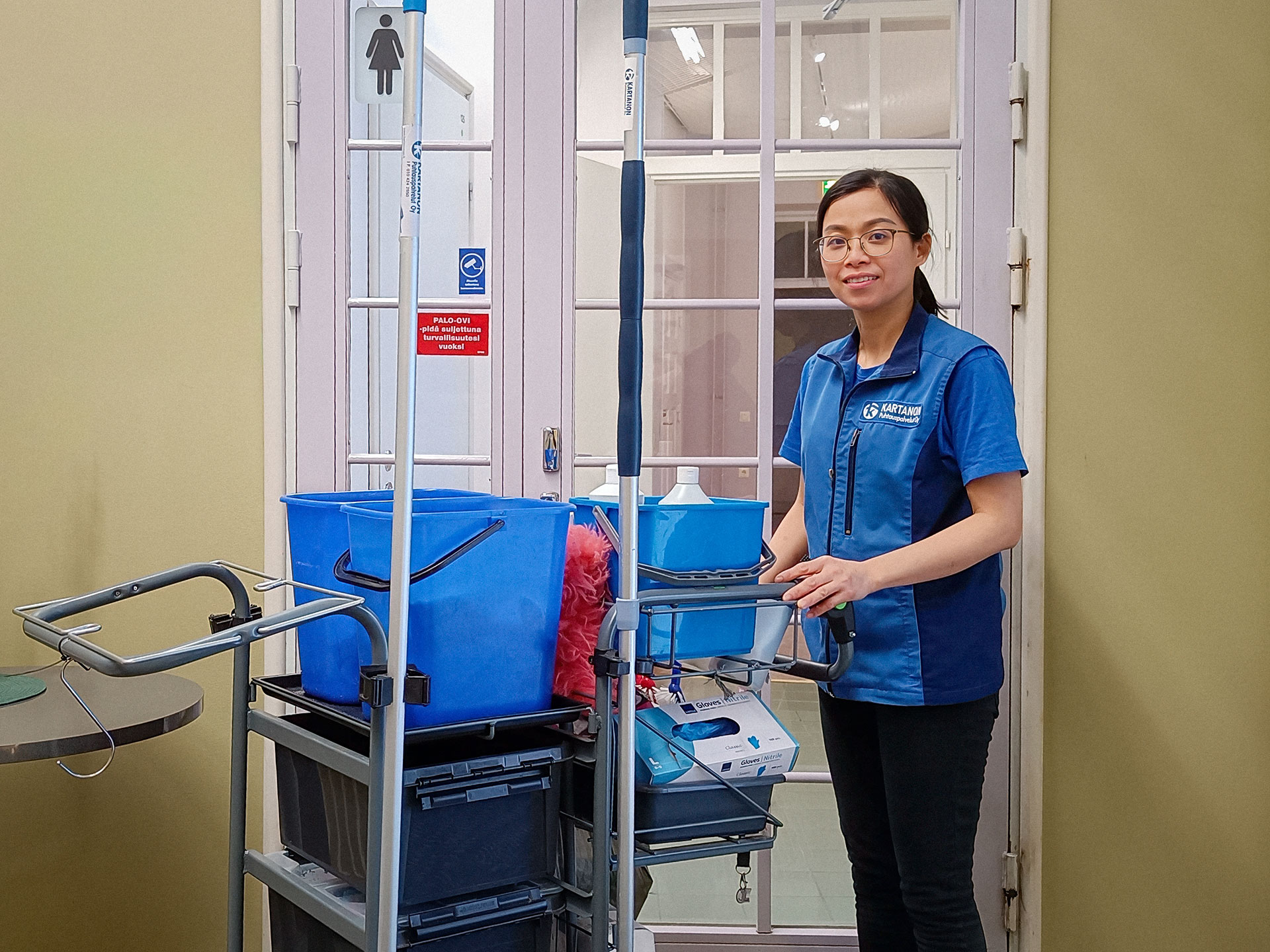 Sinisiin pukeutunut Loan siivouskärryn kanssa.