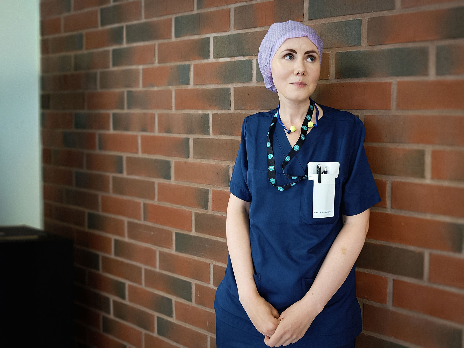 Jenita nojaa tiiliseinään, katse ylhäällä. Yllään välinehuoltajan työasu.