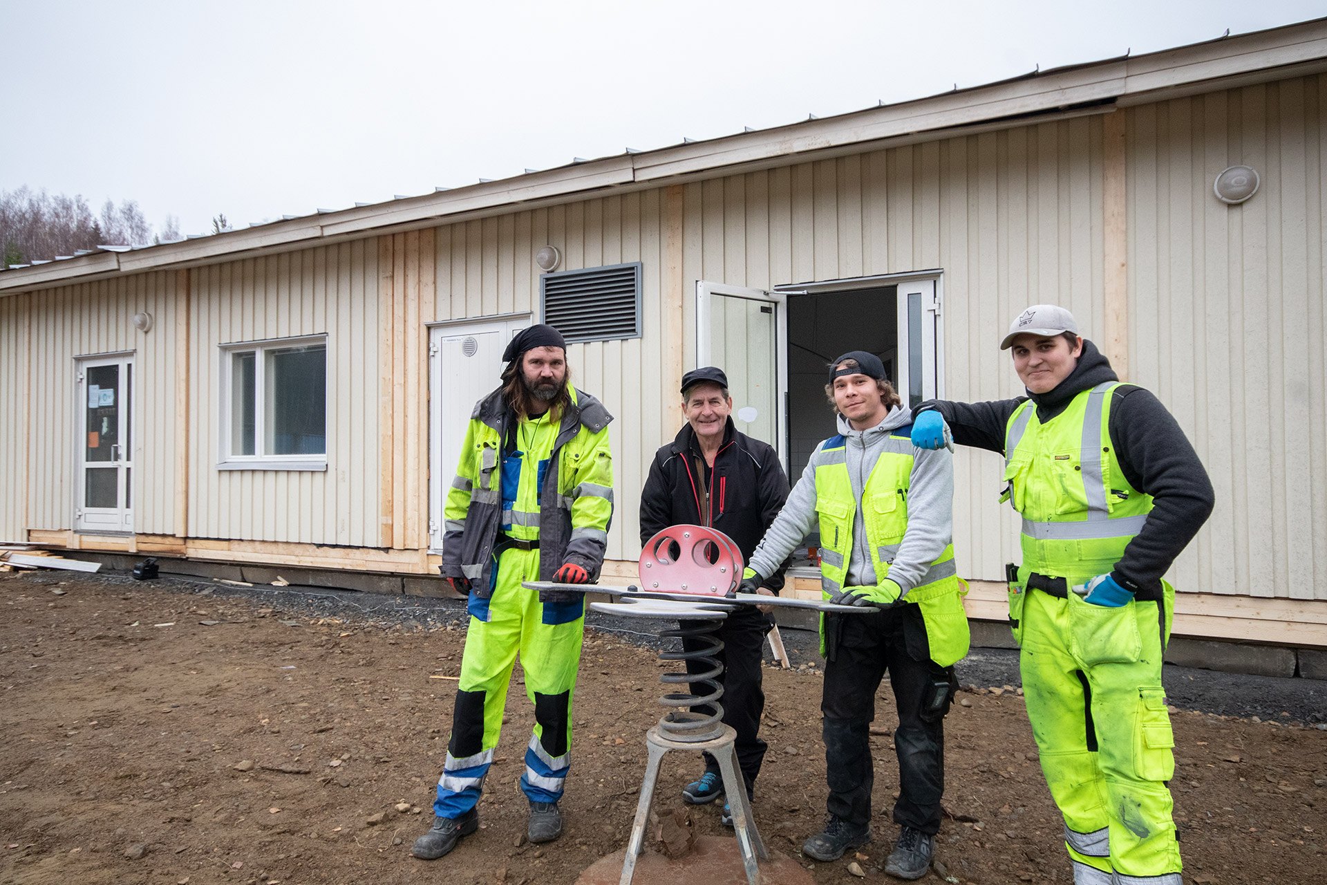 Opiskelijat seisovat päiväkotirakennuksen edessä, päällään työhaalarit. Mukana työpaikkaohjaaja. He nojaavat vieterikiikkuun.