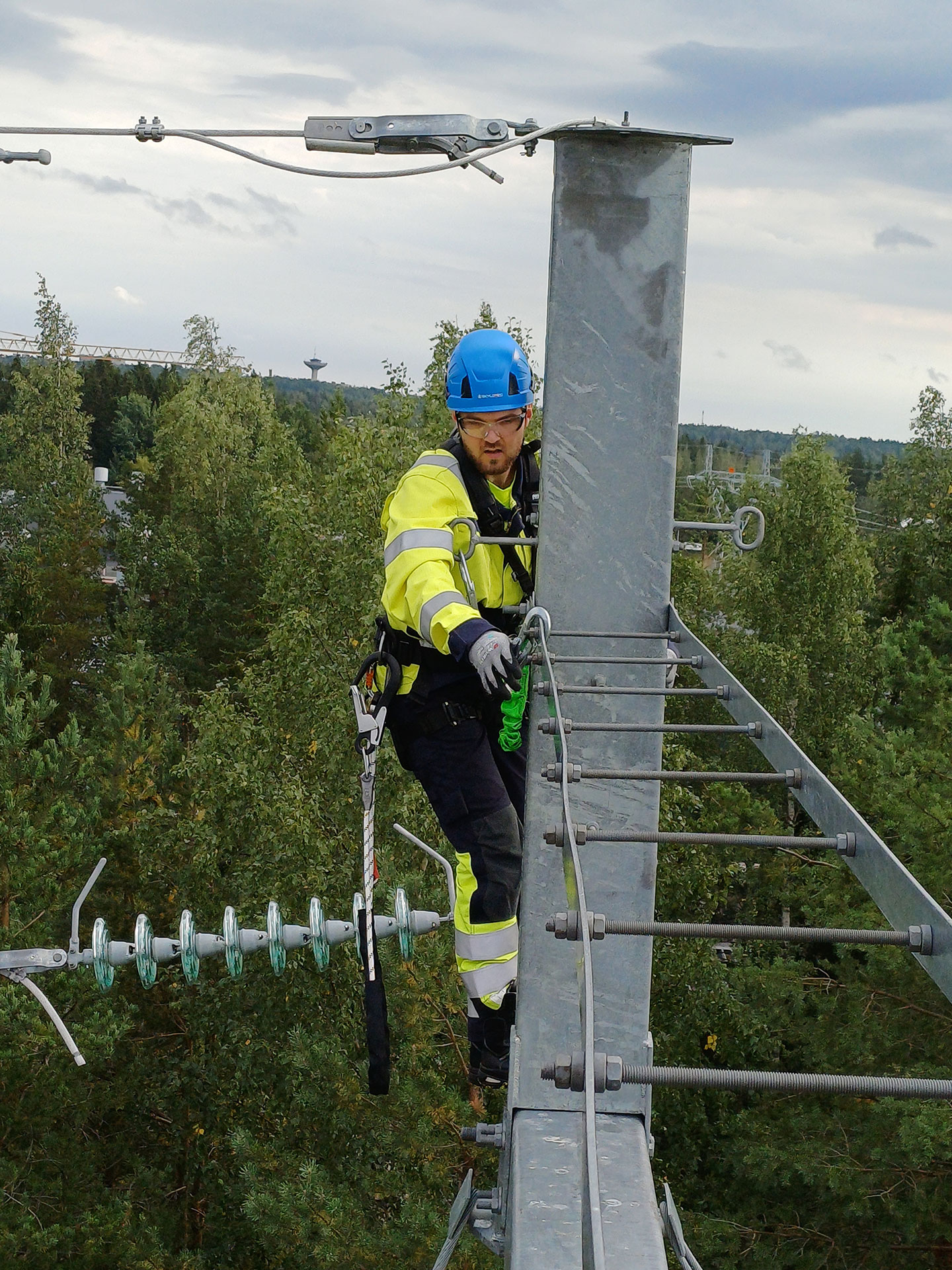 Keltamustaan haalariin ja siniseen kypärään pukeutunut Lauri kiipeilee sähköpylväissä, taustalla lehtimetsää.