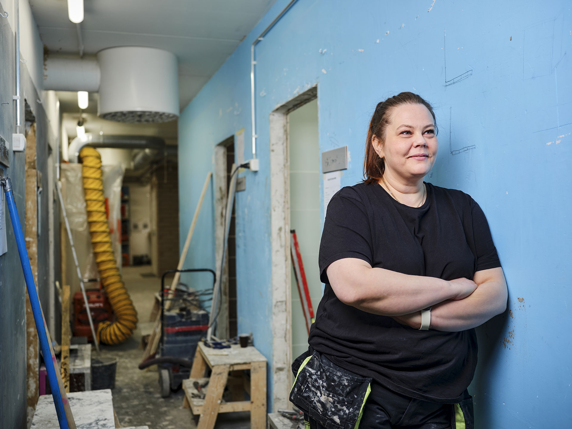 Laatoittajaksi opiskeleva Leea Perä nojaa kädet puuskassa turkoosiin seinään, taustalla käytävällä näkyy työvälineitä.