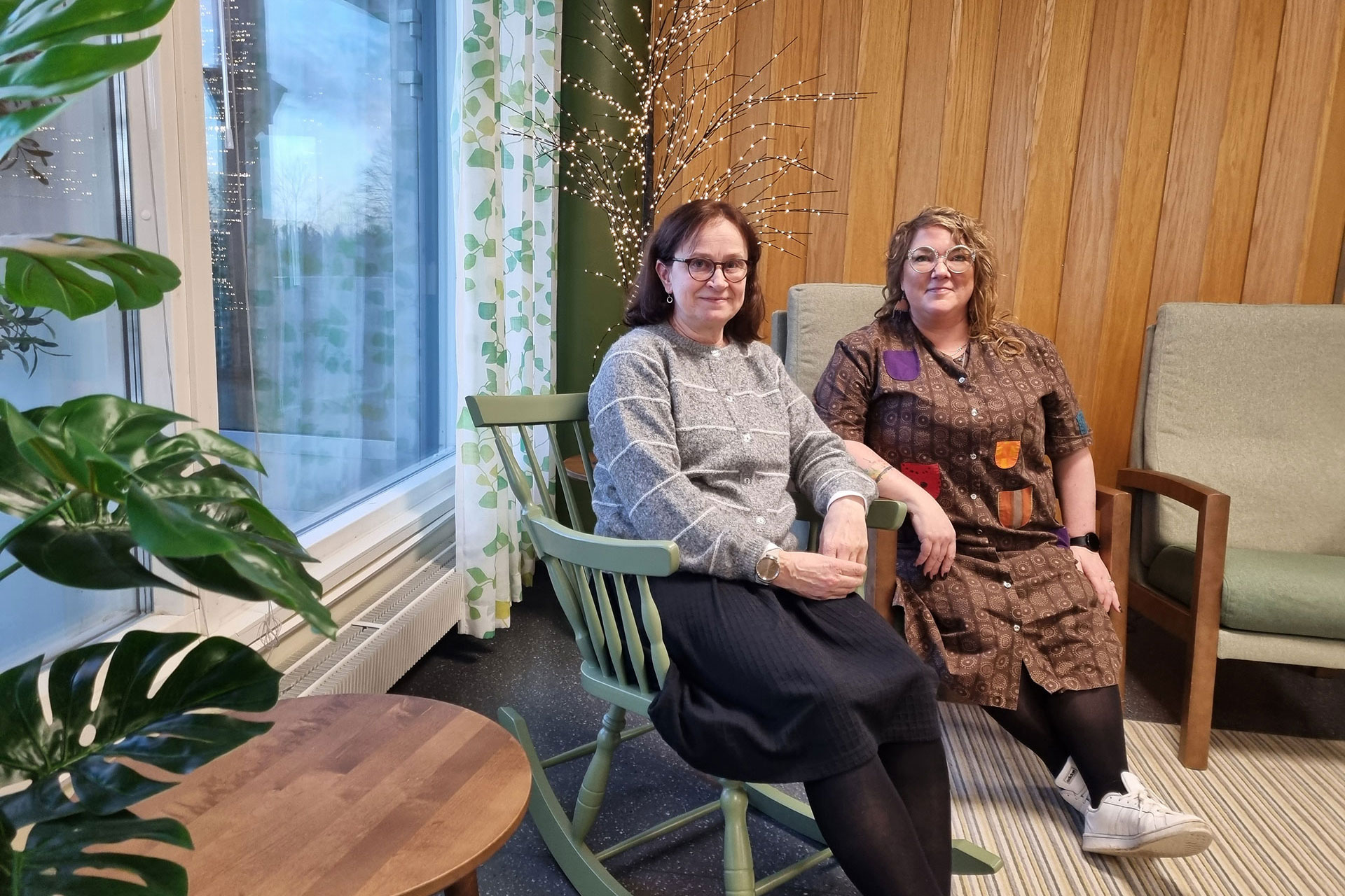 Kaksi henkilöä istuu ikkunan ääressä jouluvalopuun edustalla, toinen nojatuolissa ja toinen kiikkutuolissa.