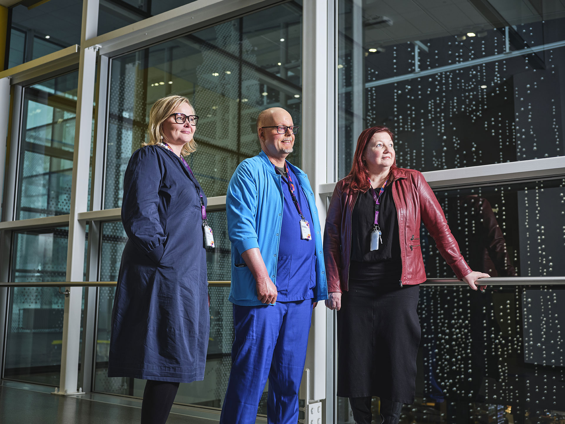 Kolme ihmistä seisoo sisällä suuren lasi-ikkunan edustalla Tampereen yliopistollisessa sairaalassa, katsovat yläviistoon.